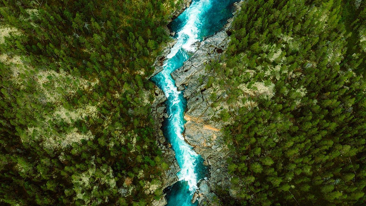 Drone high-angle photo of turquoise-colored mountain river flowing in the pine woodland with a view of the mountain peaks